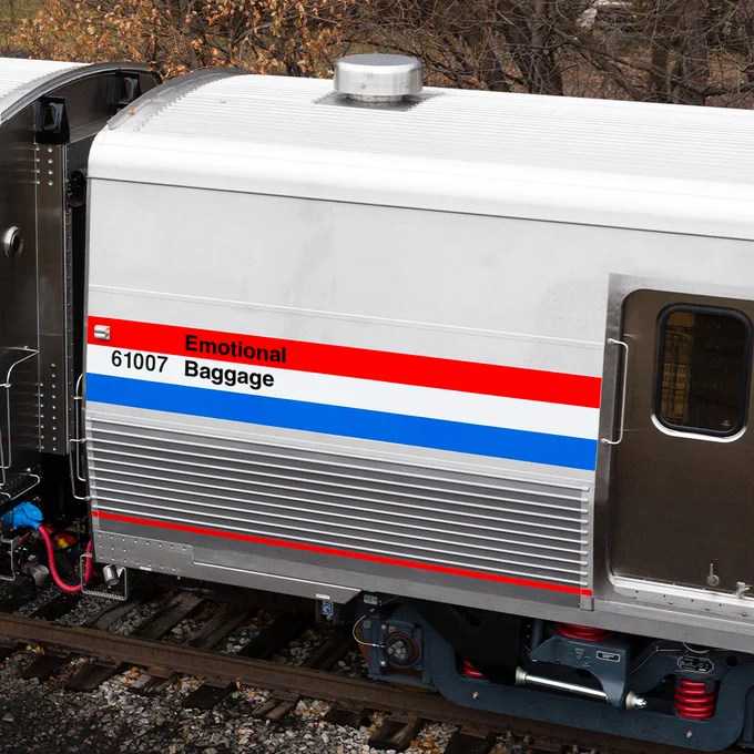Close up of the Baggage Car. The locomotive is silver with a red stripe, white stripe and blue stripe stacked in the center and running down the length of the car. The words 'emotional baggage' are written on the red and white lines. 