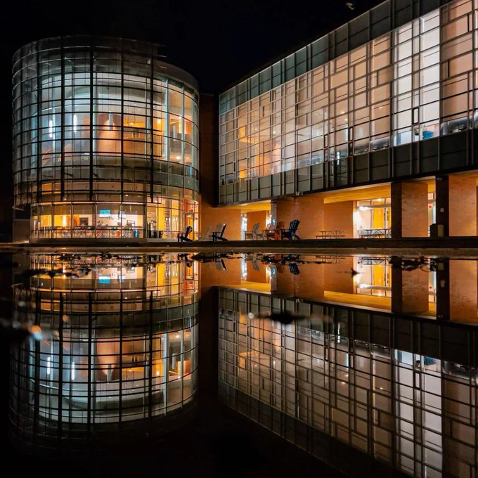 The north oshawa library at night. The sky is dark but the building is glowing and its reflection is mirrored in the reflecting pool in front of it.
