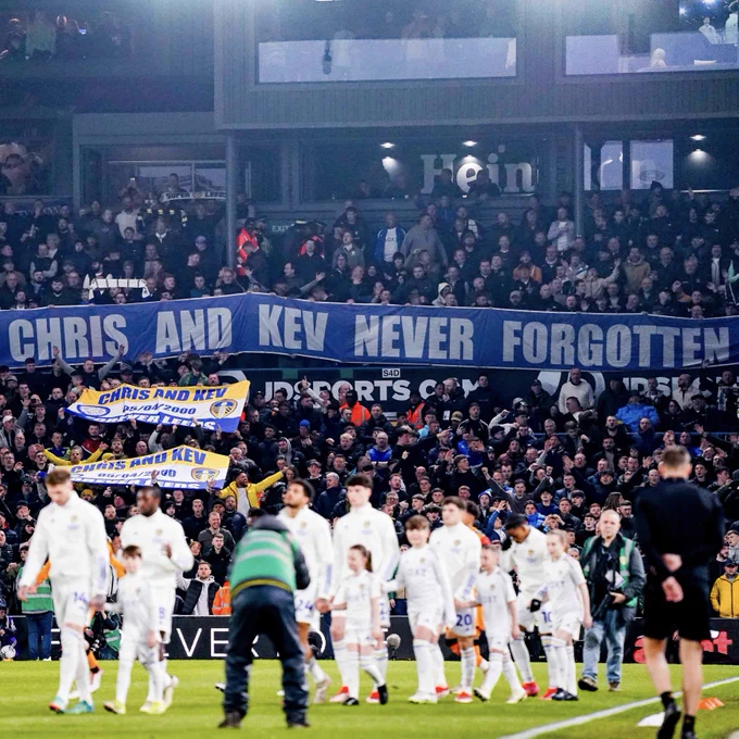 The Leeds players walk out in front of a banner which reads “RIP Chris and Kev. Never Forgotten”
