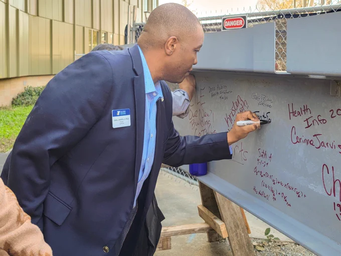 A man in a suite with a name tag that says Ivan L Harrell, II, Ph.D, President, signs a steel beam with a Sharpie pen. 