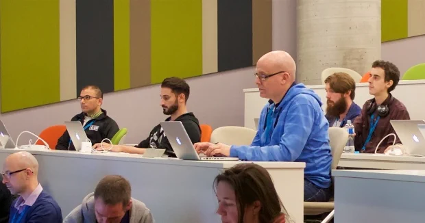 A group of people sit in a lecture theatre, listening to a presentation. Some are typing on laptops, and some are looking up at the speaker, who is not in frame.