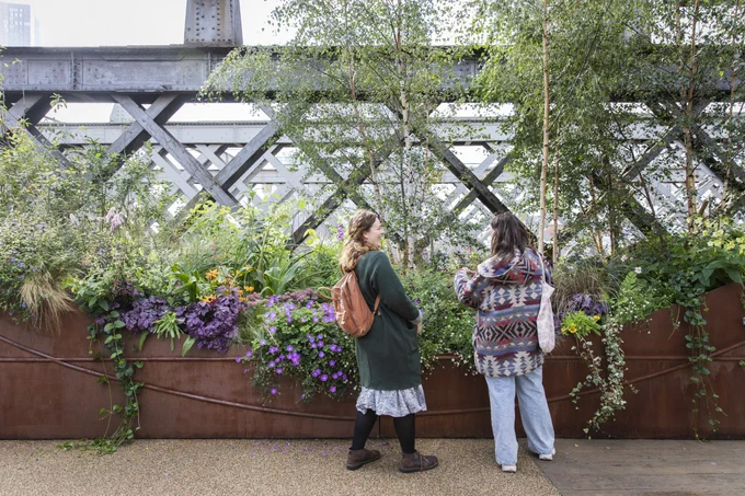 Two women chatting by a large metal planter full of flowers and plants