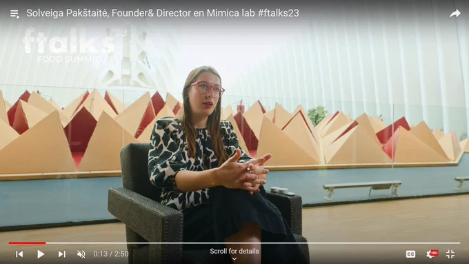 Solveiga is being interviewed in a brightly lit room at the F-talks food summit. She's wearing her signature red frame glasses and a black and white blouse in a geometric shape pattern.