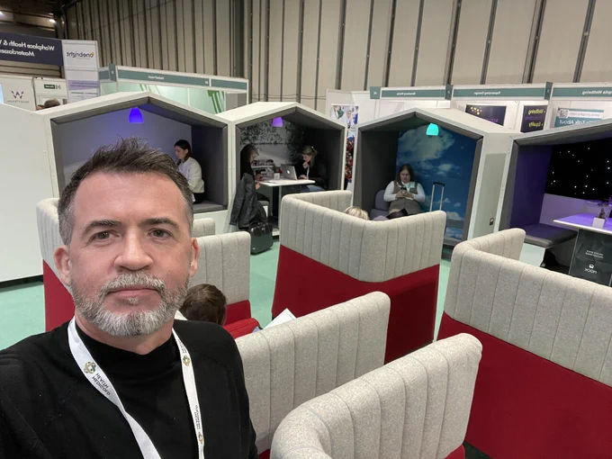Photo of David at the Health and Wellbeing Show. He's wearing a black t-shirt and behind him are various Nook pods with people relaxing or working inside them. 