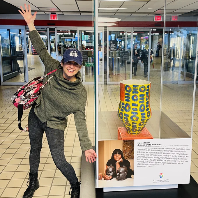 AEC’s director dressed for travel spreads her arms wide to highlight a display case featuring art by Marce Nixon-Washington at PIT.