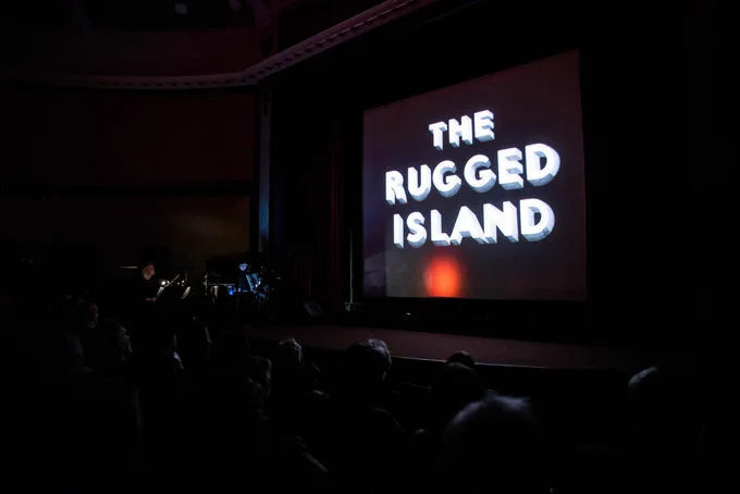 Text reading THE RUGGED ISLAND is visible on the big screen in a classic cinema interior.