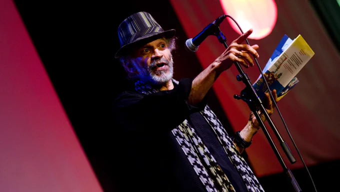 A person in a black and grey hat with a grey beard standing at a microphone with a book in one hand. They are pointing their other hand towards the audience and look like they are giving an animated reading.