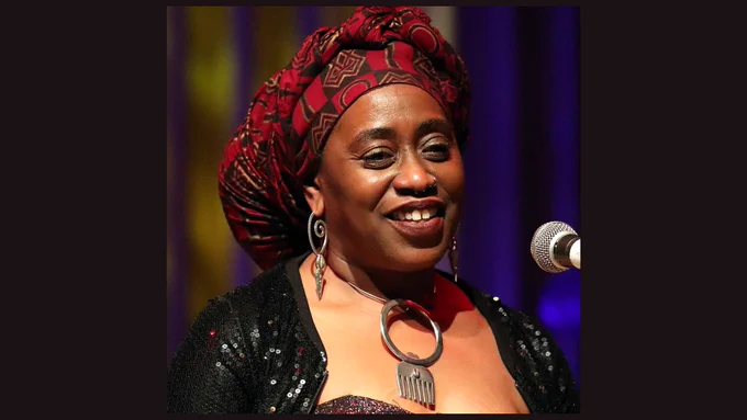A person wearing a hair scarf in rich red tones and eye-catching silver jewellery standing at a microphone. They are smiling.