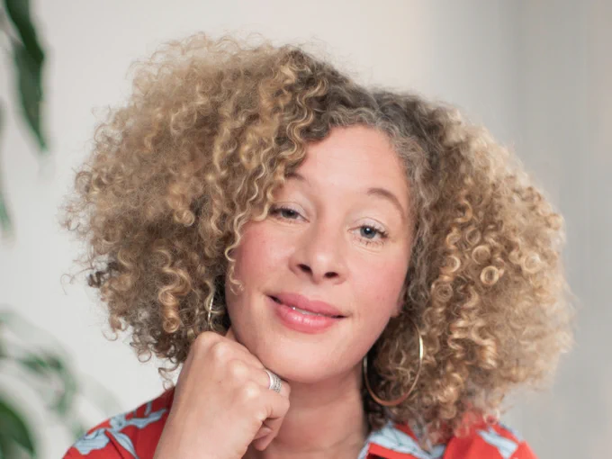 A person with blonde curly hair and silver hoop earrings smiling at the camera while resting their chin on their fist. They are smiling. 