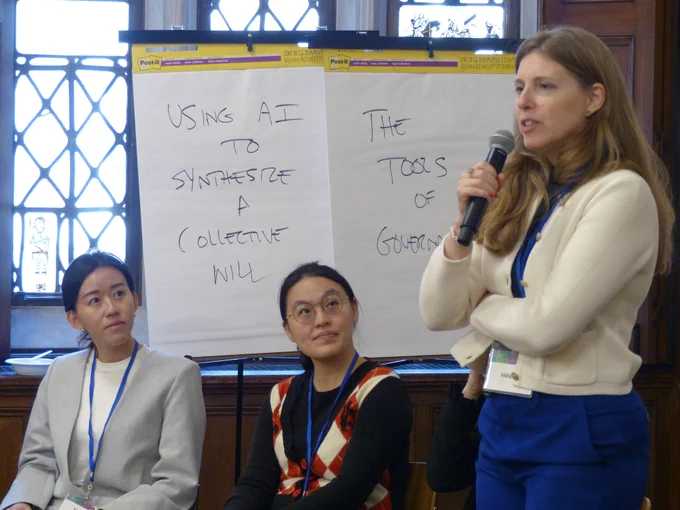 Helene Landemore stands to speak into a microphone in a conference room as attendees listen