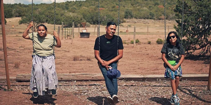 Two children and an adult sit on three swings in a dry landscape with trees in the background.