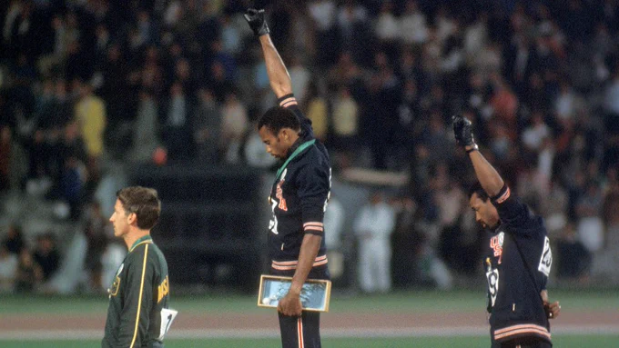 Two Black athletes raise their fists in protest at the 1968 Olympics while a white athlete faces away from them. 