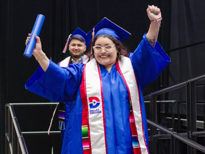 A female graduate, with her degree in hand, walks down the stage ramp with her arms raised to the sky. 