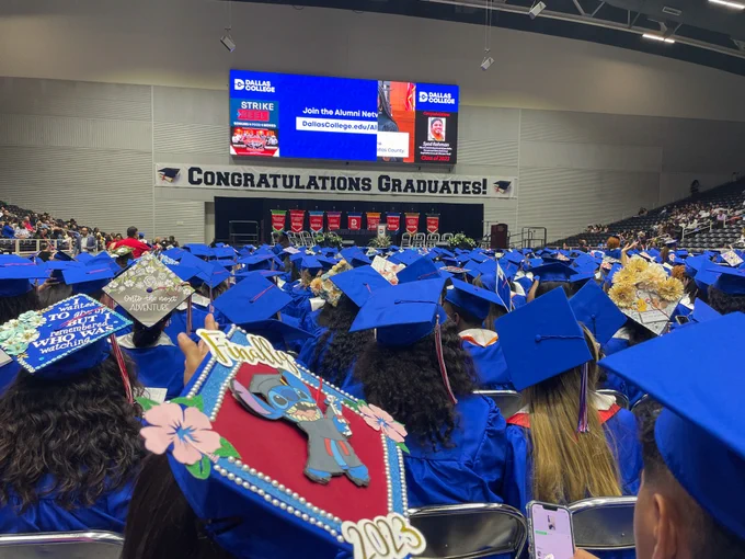 View from the back of the graduate row as we look onward toward the front of the stage. A large banner at the stage reads CONGRATULATIONS GRADUATES