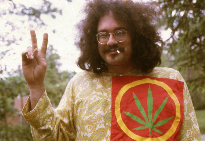 A mustachioed, wild-haired, and smiling John Sinclair flashes the peace sign while smoking a cigarette and looking into the camera. Photo by Leni Sinclair in 1968.