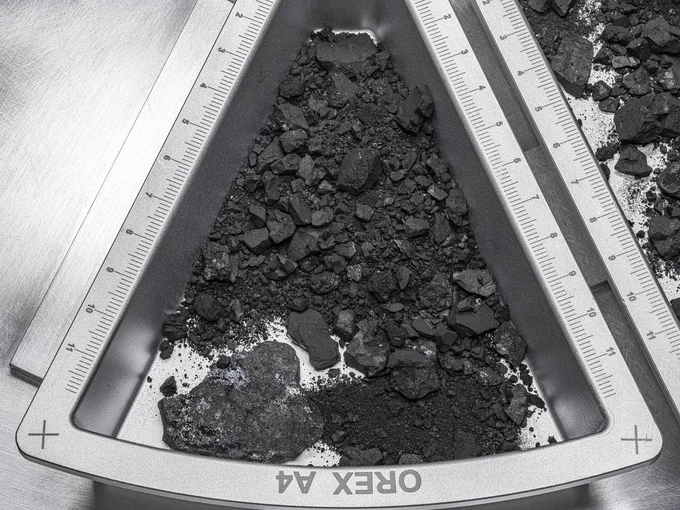 Silver wedge-shaped container has a ruler on the sides in centimeters. Small dark rocks and rubble are within the container, which says OREX A4. 