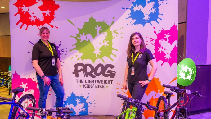 Kirsteen and Alice from Frog Bikes are standing at their stall from the Bikeability Conference. They are posing with a range of Frog cycles in front of a large branded backdrop. There is a green Frog Easter Egg peaking out from behind it!
