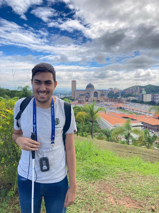 Estou sorridente em um jardim do Santuário Nacional de Nossa Senhora aparecida, com vista panorâmica para o centro de peregrinação. ao redor do Santuário, o Centro de Apoio ao Romeiro e parte da cidade de Aparecida.

Uso camiseta branca, calça jeans, estou com mochila nas costas e um rádio receptor com fones de ouvido pendurado no pescoço. Seguro uma bengala branca. O céu está azul com algumas nuvens.