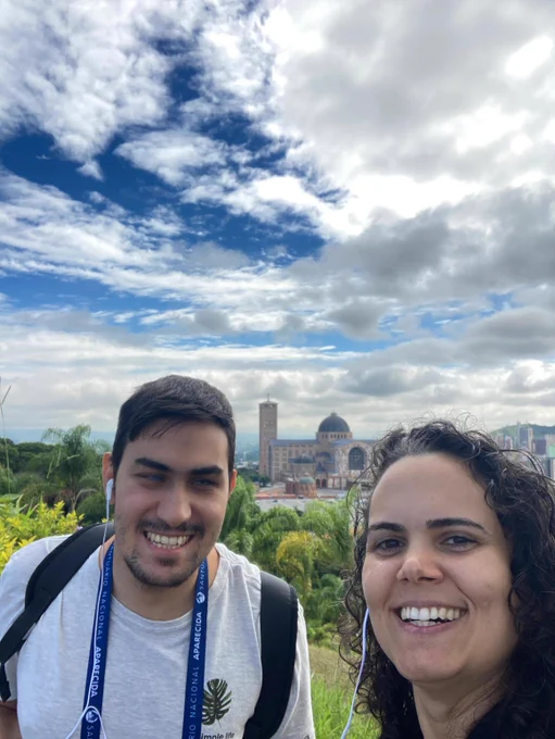 Selfie de Flávia Machado comigo jardim do Santuário Nacional de Nossa Senhora aparecida, no mesmo momento e cenário da foto anterior. Ela é uma mulher branca de cabelo cacheado e usa uma camisa preta. Estamos sorrindo.

