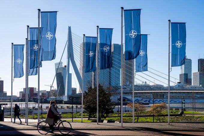 NAVO-vlaggen hangen nabij de Erasmusbrug in Rotterdam (te zien op de achtergrond). Een vrouw fietst op de voorgrond voorbij. Het is zonnig weer met een blauwe lucht. De foto is vorig gemaakt op 4 april, in het kader van 'NAVO-dag'.