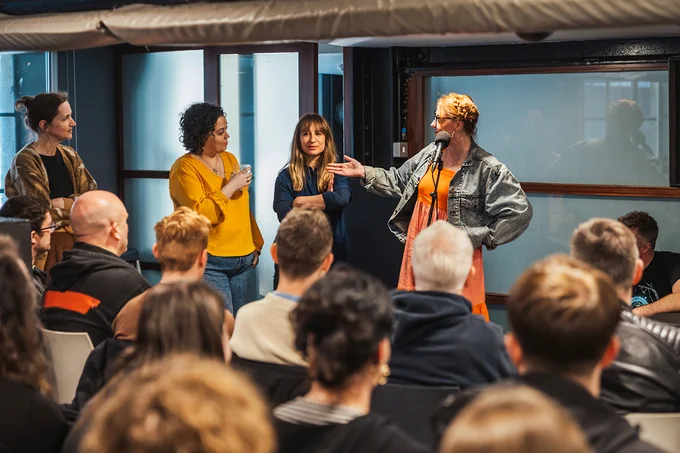 Four panellists stand in front of a seated audience at a networking event. One panellist is stood in front of a microphone and is gesturing towards the rest of the panel.
