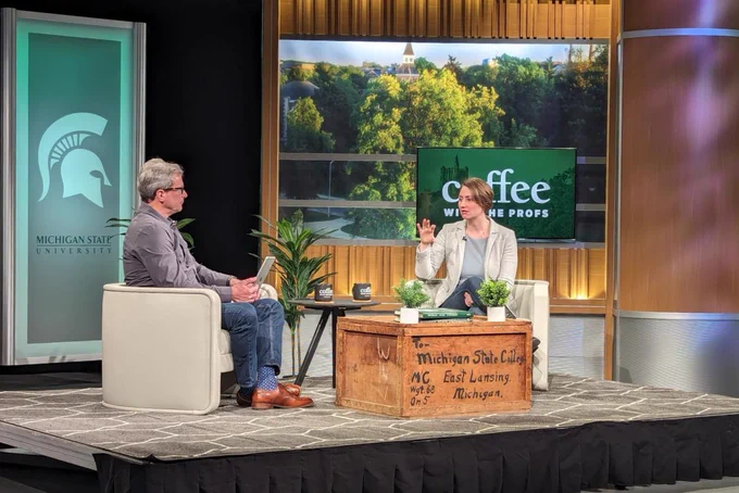 Two people sit in chairs on a stage. In front of them is a wooden packing crate that acts as a coffee table. The packing crate says: “To: Michigan State College, East Lansing, Michigan.” Behind the two people is a Michigan State University banner with the Spartan helmet logo. Another screen shows a campus photo of trees with the point of a building tower poking through and the text: “Coffee with the Profs.”