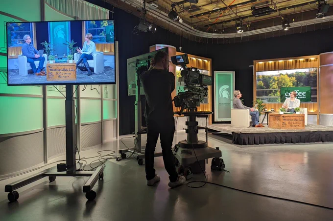 A TV monitor shows two people sit in chairs on a stage for a behind the scenes look of the MSU Coffee with the Profs filming. A person stands behind a large video camera on wheels as they watch and listen to the conversation between two people on the stage.