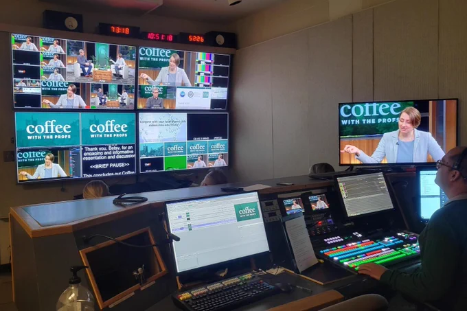 A production room with four TV monitors shows different parts of the Coffee with the Profs filming, including the two people talking on stage, the live transcript of what the people are saying, and the Coffee with the Profs logo. A person sits behind a desk with three computers and an audio board to help mix the sound during the filming.