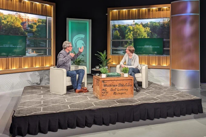 Two people sit in chairs on a stage and have an animated conversation. In front of them is a wooden packing crate that acts as a coffee table. The packing crate says: “To: Michigan State College, East Lansing, Michigan.” Behind the two people is a Michigan State University banner with the Spartan helmet logo. Another screen shows a campus photo of trees with the point of a building tower poking through and the text: “Coffee with the Profs.”