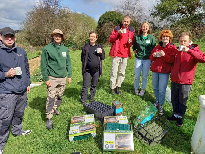 People from the Green Communities project and Devon Wildlife Trust's Saving Devon's Treescapes project stood outside in the sunshine, holding cups of tea, next to the tree nursery kits.