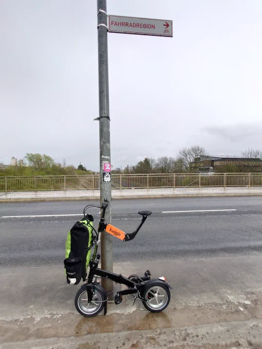 Kwiggle folding bike with pannier on front rack, leaning against a sign "bicycle region"