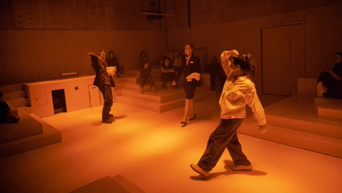 In a room lined with step-like blocks that jut out from the walls, three people move in different directions. A white woman with tied-back blonde hair strides forward purposefully, a towel over her arm; a Caucasian man with sandy hair holds a bucket and has his arm back over his shoulder, as if about to throw something; a small, dark haired woman facing opposite him is doing the same motion.
