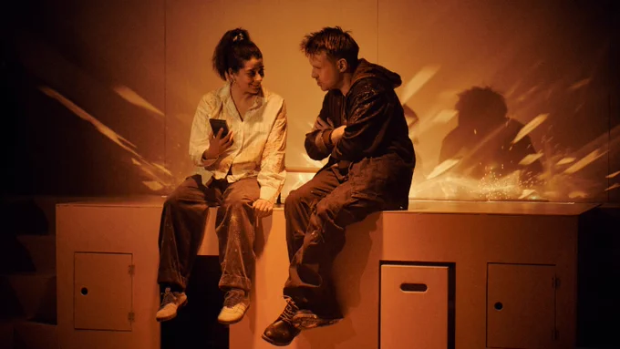 A small, dark haired woman with mid length hair in a ponytail and a Caucasian man with sandy hair and blue eyes sit on a cabinet built into an orange wall. They are both covered in custard. The woman holds up a small block-shaped object to show to the man while he speaks.