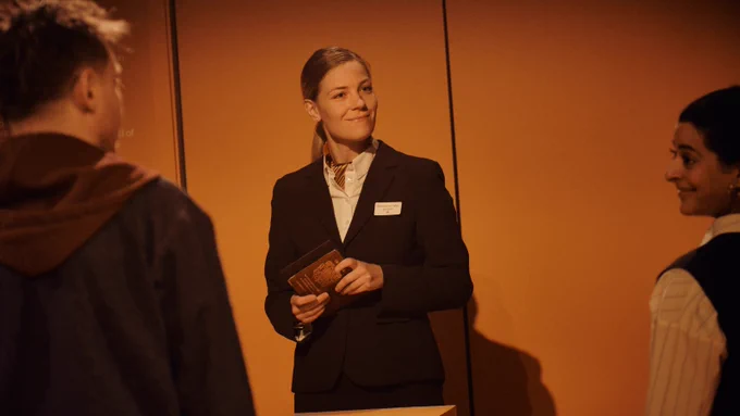 A white woman with tied-back blonde hair, wearing a dark suit, stands in front of two people who are facing away from the camera. She is very neat, with a neckscarf, and holds open a passport.