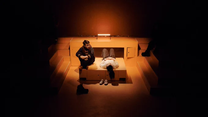 In a room lined with step-like blocks that jut out from the walls, a Caucasian man with sandy hair sits on a freestanding box on wheels with his knees pulled up around him. Next to him on the box lies a small, dark haired woman. Two pairs of shoes are on the ground next to them. The room is lit by one orange fluorescent bar that glows.