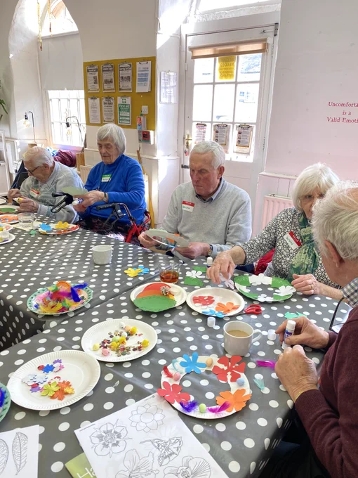 A group of older people sit around tables covered with spotty table cloths sticking paper flowers to make spring wreaths