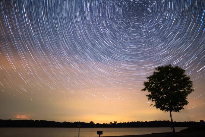 A circular star trail is seen in a purple and yellow night sky. In the foreground, you can see a silhouette of a tree. 