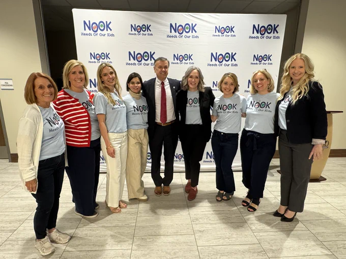 8 women posing with a man in the middle in front of a large screen that says The NOOK.