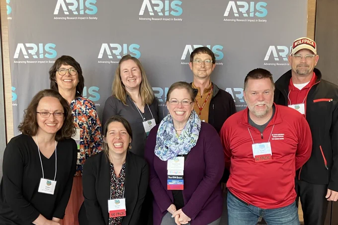 8 smiling people posing in front of a gray background with ARIS written on it.