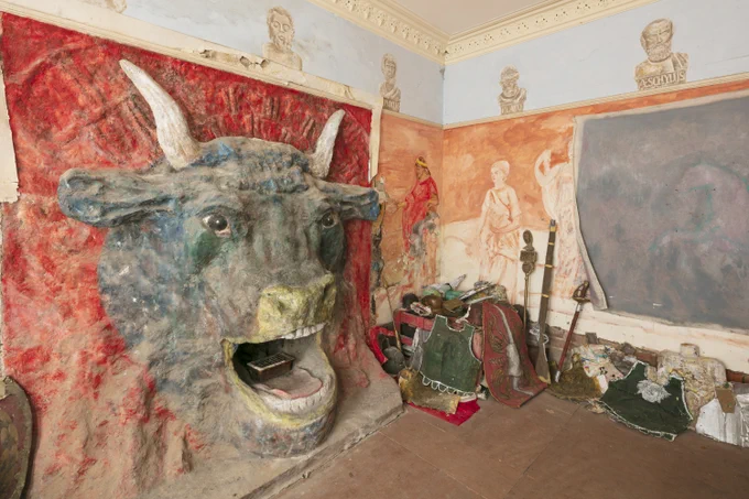 A photograph of a ground floor room with a large Minotaur head surrounding a fireplace and portraits of Greek philosophers. Artwork by Ron Gittins.
