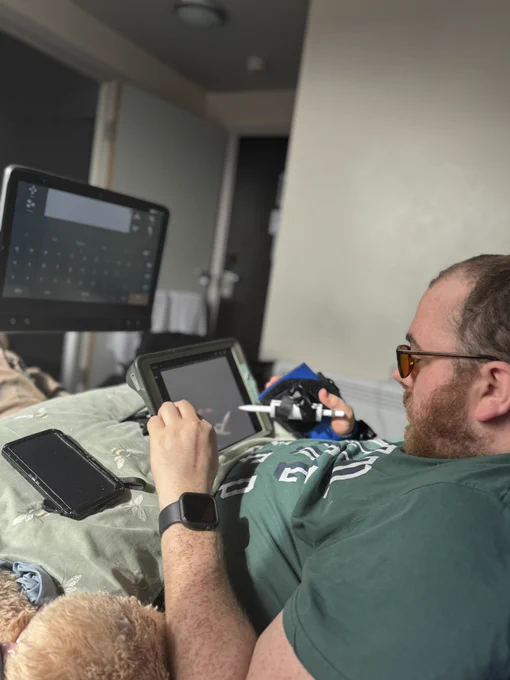 Side profile of me a white non binary trans man wearing a green t shirt with brown hair and a beard. Holding an Apple Pencil using a grip aid and drawing on an iPad. There’s an eye gaze device on the far left positioned above the iPad and on a stand