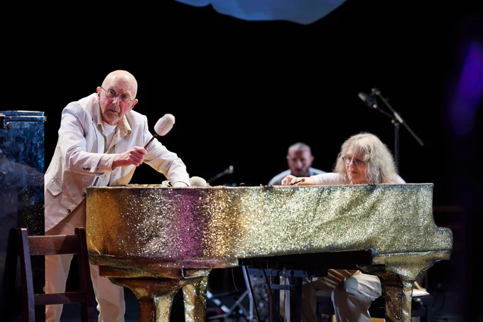 A person in a white suit standing next to a golden, sparkly piano.
