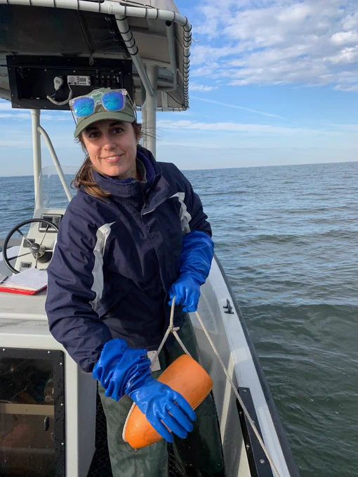 A young female scientist stands on a boat, wearing blue gloves and holding an orange float