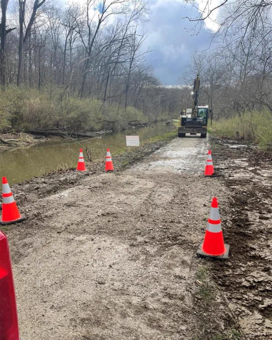 View down an unpaved trail lined with safety cones. On the left, water fills the canal along the path. Heavy equipment operates in the distance.