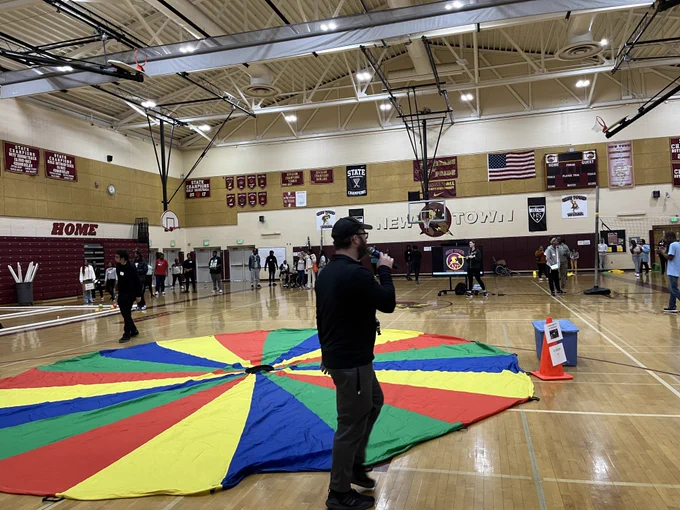Photo of a person in a school gym next to a colorful parachute. Other people are standing around the gym.