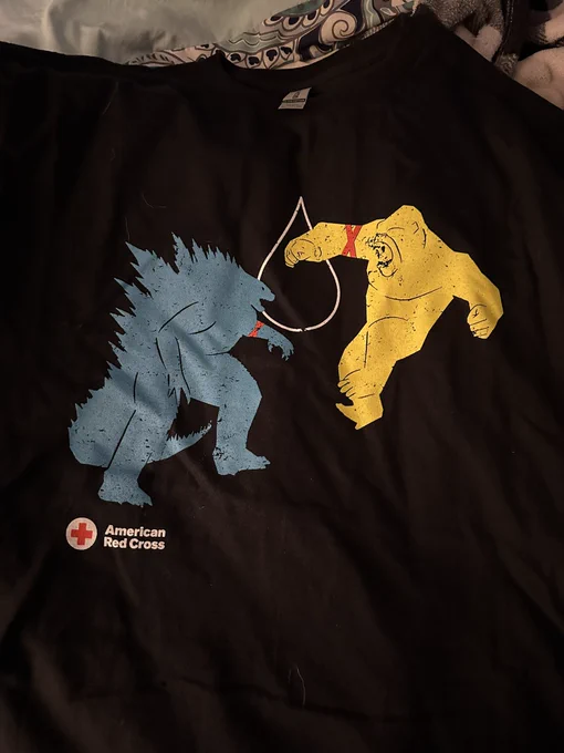 Black T-shirt with image of Godzilla fighting Kong and both monsters have red tape wrapped around an arm as if they had recently donated blood. And the American Red Cross logo is in the bottom left corner.