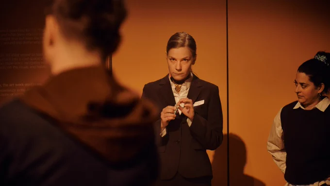 A white woman with tied-back blonde hair, wearing a dark suit, stands next to a small, dark-haired woman with mid-length hair tied back into a ponytail. The woman in the dark suit holds something up to her finger. In the foreground of the image, slightly out of focus, is a Caucasian man with sandy hair.