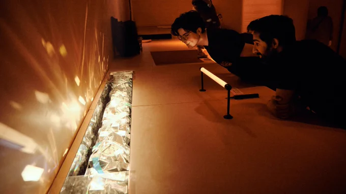 A long, thin lighting fixture emits reflections and refractions of light against an orange wall; two audience members look at it, smiling.