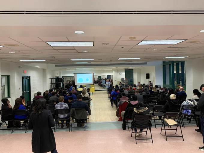 The photo shows a room with a screen displaying a slideshow presentation and speaker standing at the front as well as rows of chairs filled with audience members facing forward