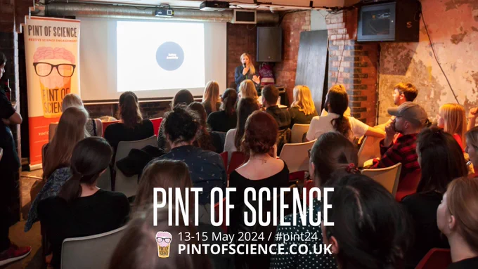 Backs of people sat on chairs, a person stands at the front with a microphone, a presentation behind them. Text on image: PINT OF SCIENCE 13-15 May 2024 / #pint24 with a link to the website.